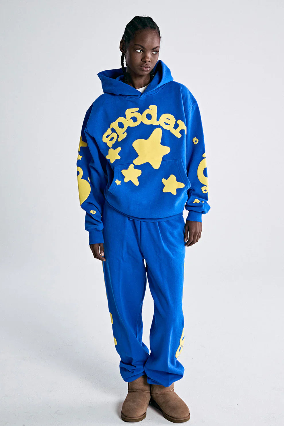 Wearing the Sp5der 440GSM Heavyweight Hoodie in royal blue with yellow star 3D puff print and matching sweatpants, this outfit serves ultimate Y2K streetwear vibes on a white background, completed with comfy brown slippers.