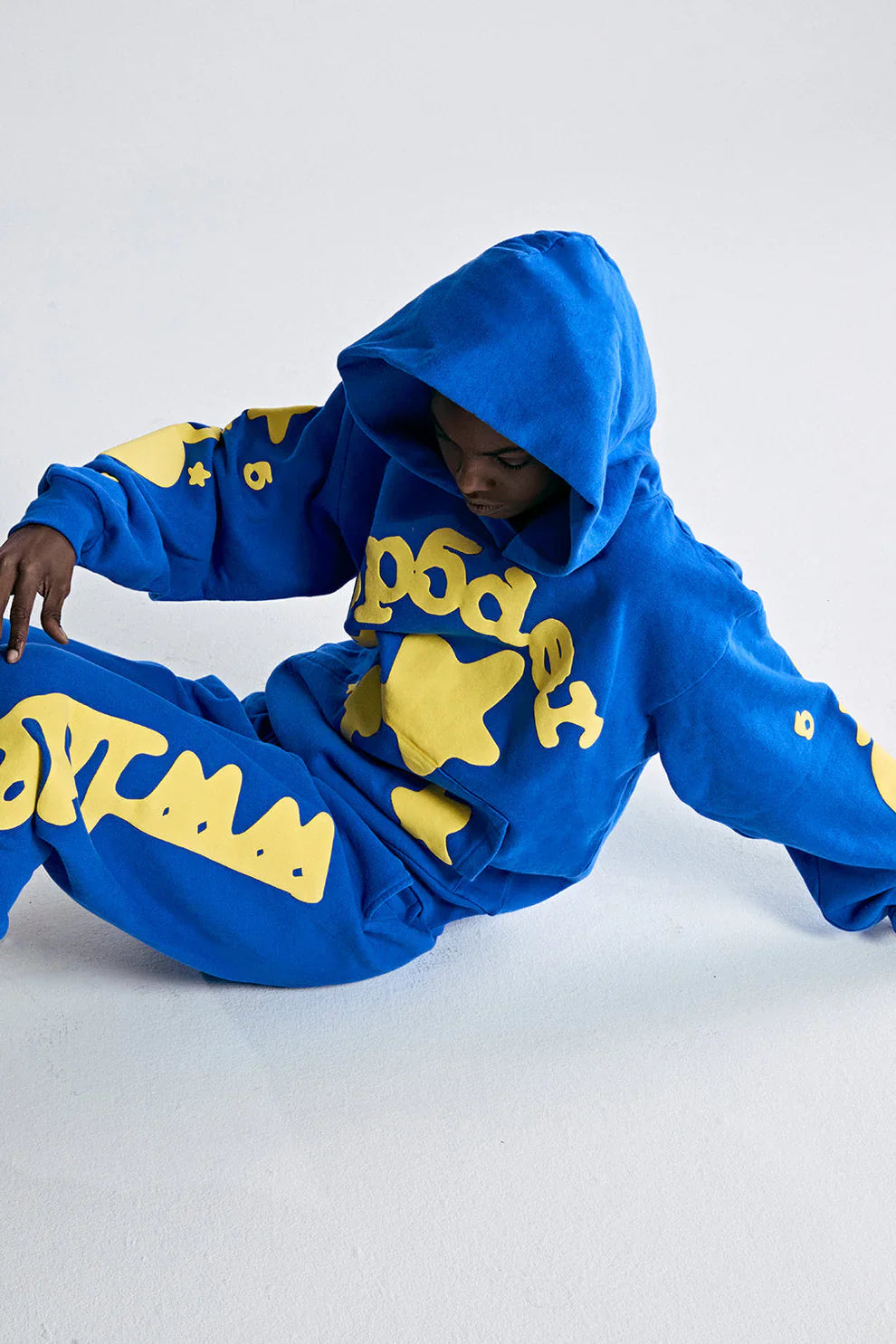 Wearing the Sp5der 440GSM Heavyweight Hoodie in royal blue with yellow star 3D puff print, a person sits on the floor against a light background, looking down—a true nod to Y2K streetwear style.