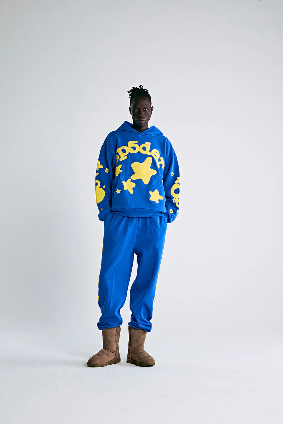 A person stands against a plain light background wearing the Sp5der 440GSM Heavyweight Hoodie in Royal Blue with Yellow Star 3D Puff Print, styled with matching pants and tan boots, channeling a true Y2K streetwear vibe.