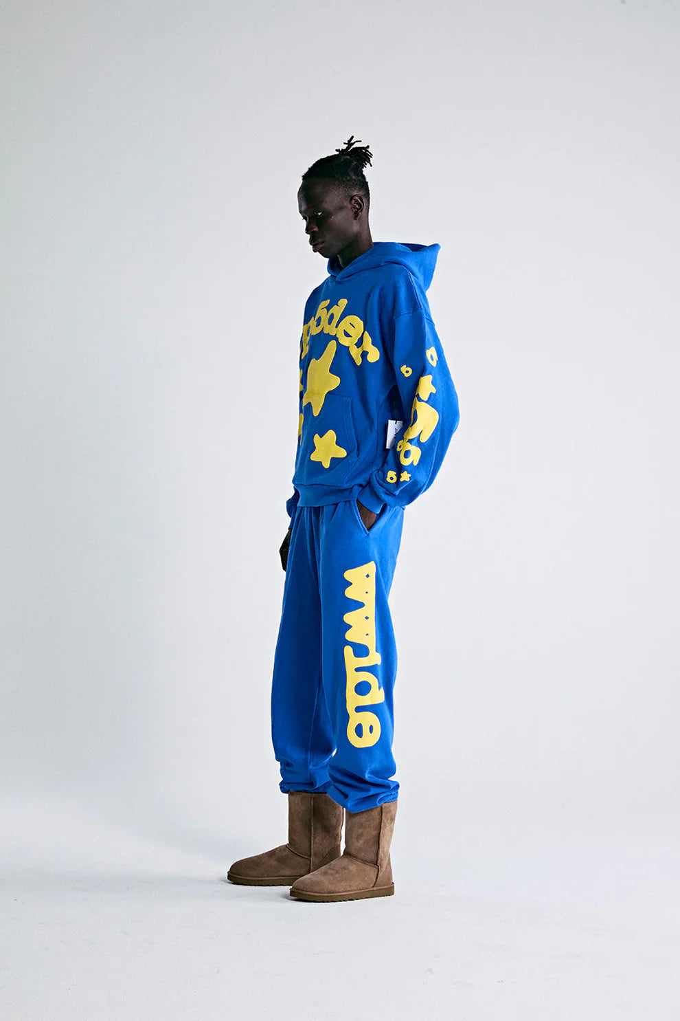 Against a plain light background, a person in profile wears the Sp5der 440GSM Heavyweight Hoodie in royal blue with yellow star 3D puff print, paired with matching sweatpants and brown boots.
