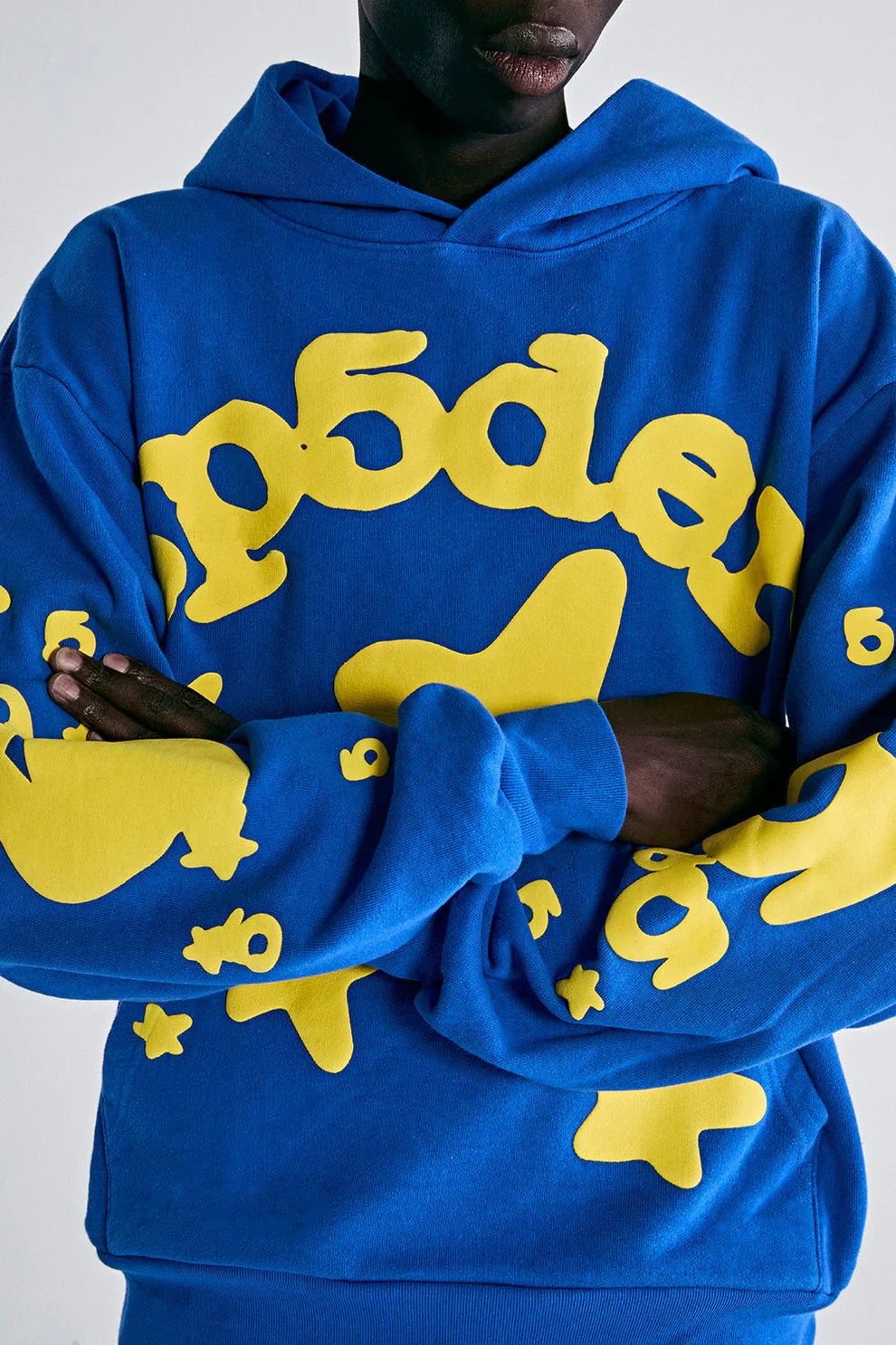 Person wearing a Sp5der 440GSM Heavyweight Hoodie – Royal Blue with Yellow Star 3D Puff Print, arms crossed, only torso and lower face visible against a plain background.