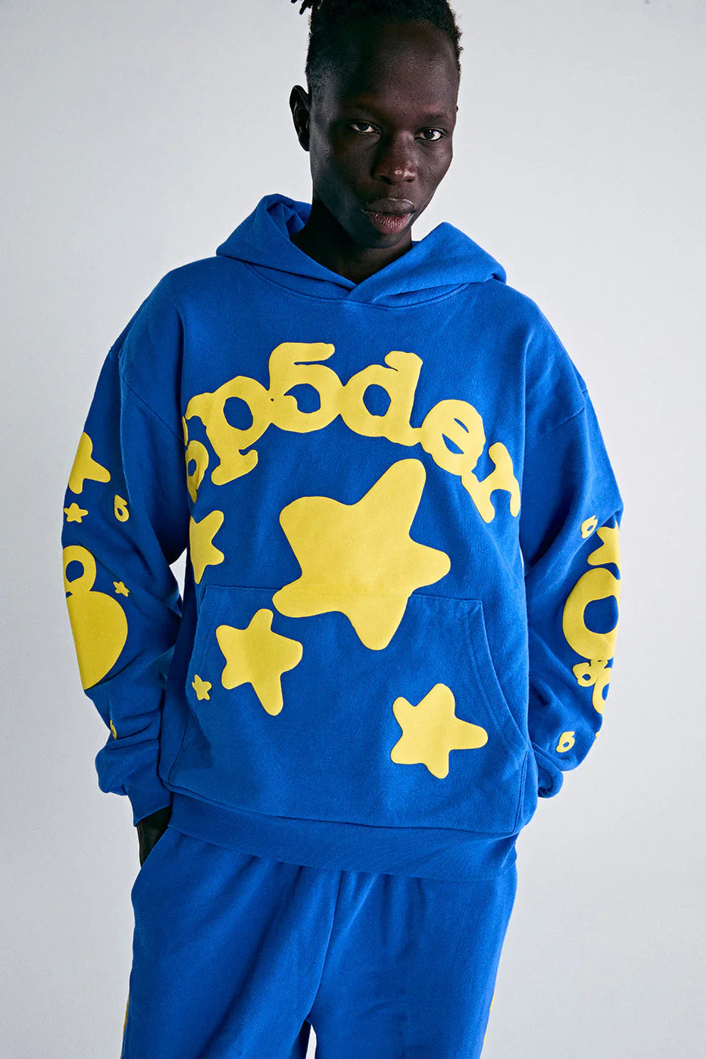 Showcasing bold Y2K streetwear vibes, a person models the Sp5der 440GSM Heavyweight Hoodie in royal blue with yellow star 3D puff print and matching pants against a plain background, highlighting the premium cotton fleece.