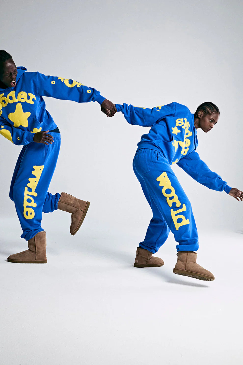 Two people wear matching Sp5der 440GSM Heavyweight Hoodies in royal blue with yellow star 3D puff print, paired with tan boots, holding hands and leaning in opposite directions against a plain background.
