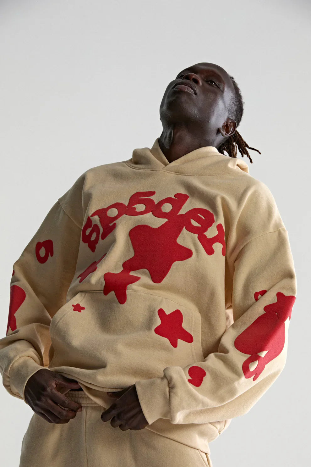 Wearing the Sp5der 440GSM Heavyweight Star Hoodie in cream with red puff print, an oversized streetwear pullover, a person stands against a plain background, channeling modern style while looking slightly upward with hands near their waist.