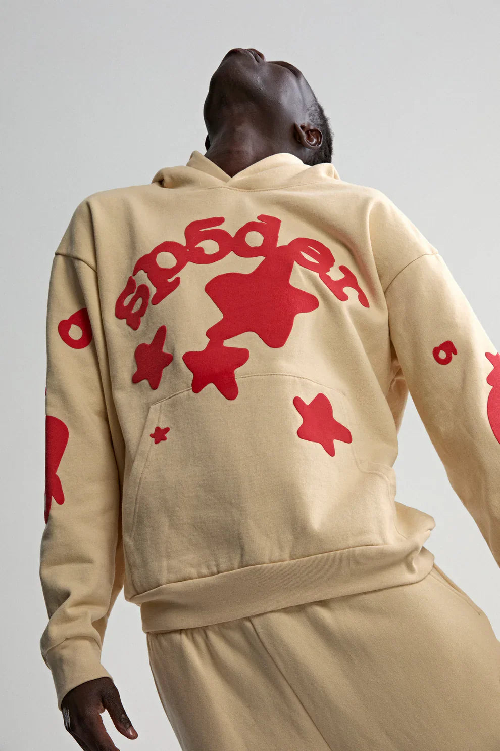 A person in a Sp5der 440GSM Heavyweight Star Hoodie – Cream with Red Puff Print stands with head tilted back. The oversized streetwear pullover's bold red stars and stylized text pop against the plain, light background.