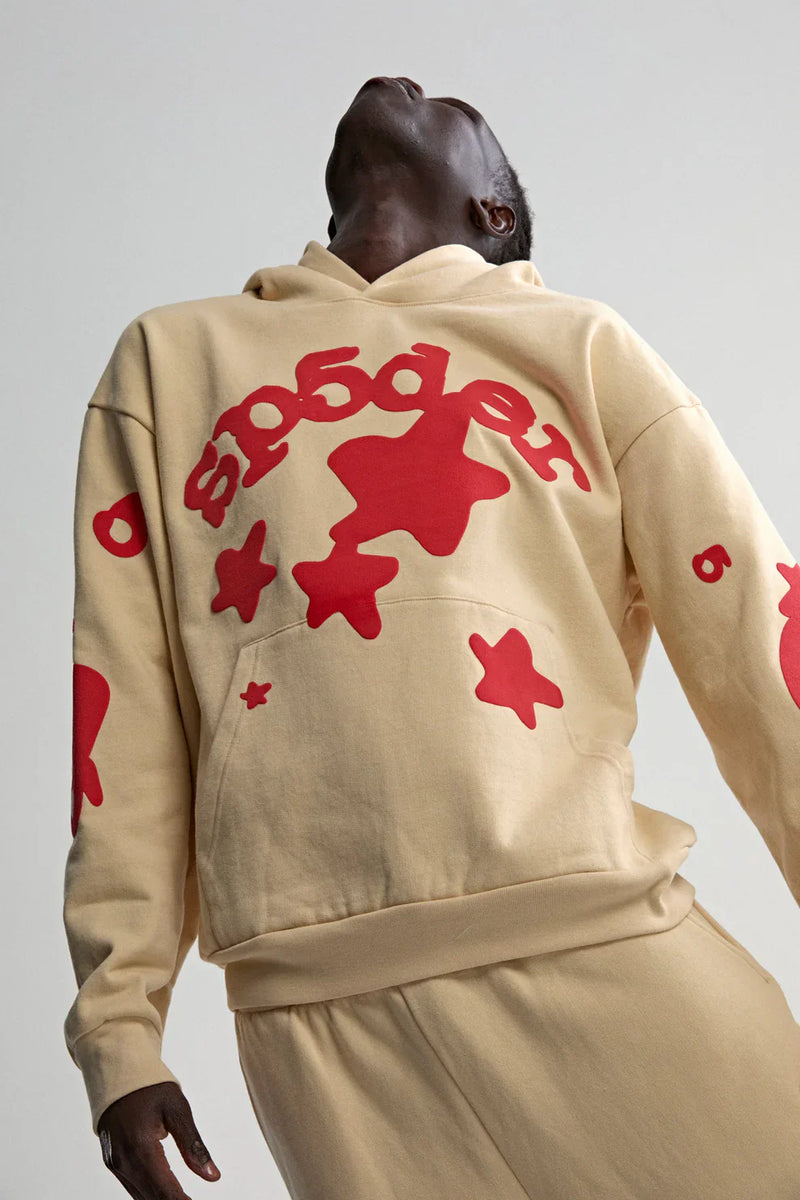 A person in a Sp5der 440GSM Heavyweight Star Hoodie – Cream with Red Puff Print stands with head tilted back. The oversized streetwear pullover's bold red stars and stylized text pop against the plain, light background.