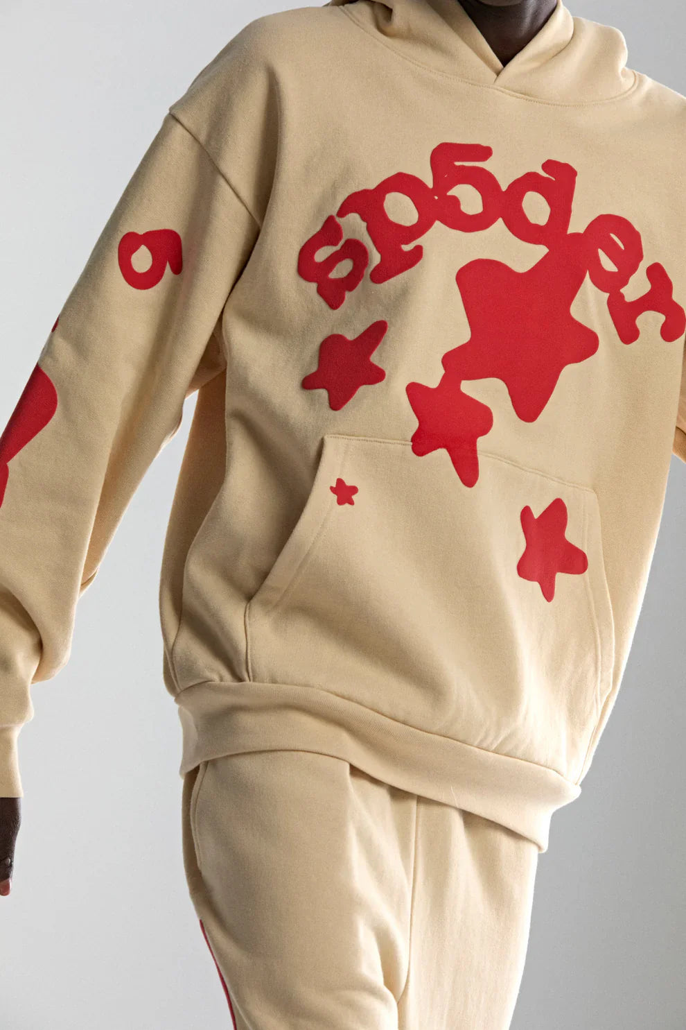 Person wears the Sp5der 440GSM Heavyweight Star Hoodie in cream with red puff print—an oversized streetwear pullover featuring bold star graphics and standout “sp5der” text, paired with matching pants for a striking look.
