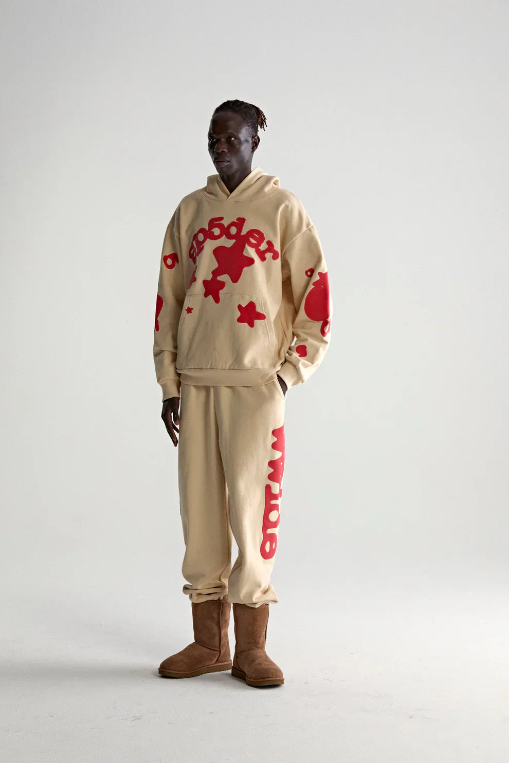 A person wears the Sp5der 440GSM Heavyweight Star Hoodie in cream with red puff print, paired with matching sweatpants and brown boots, embodying oversized streetwear style against a plain light gray background.