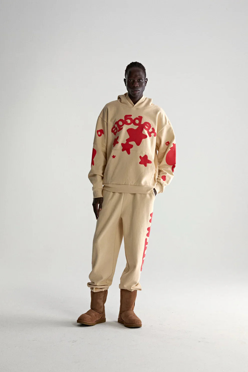 A person poses against a plain background in the Sp5der 440GSM Heavyweight Star Hoodie—cream with red puff print—styled with matching sweatpants and brown boots for an oversized, streetwear-inspired look.