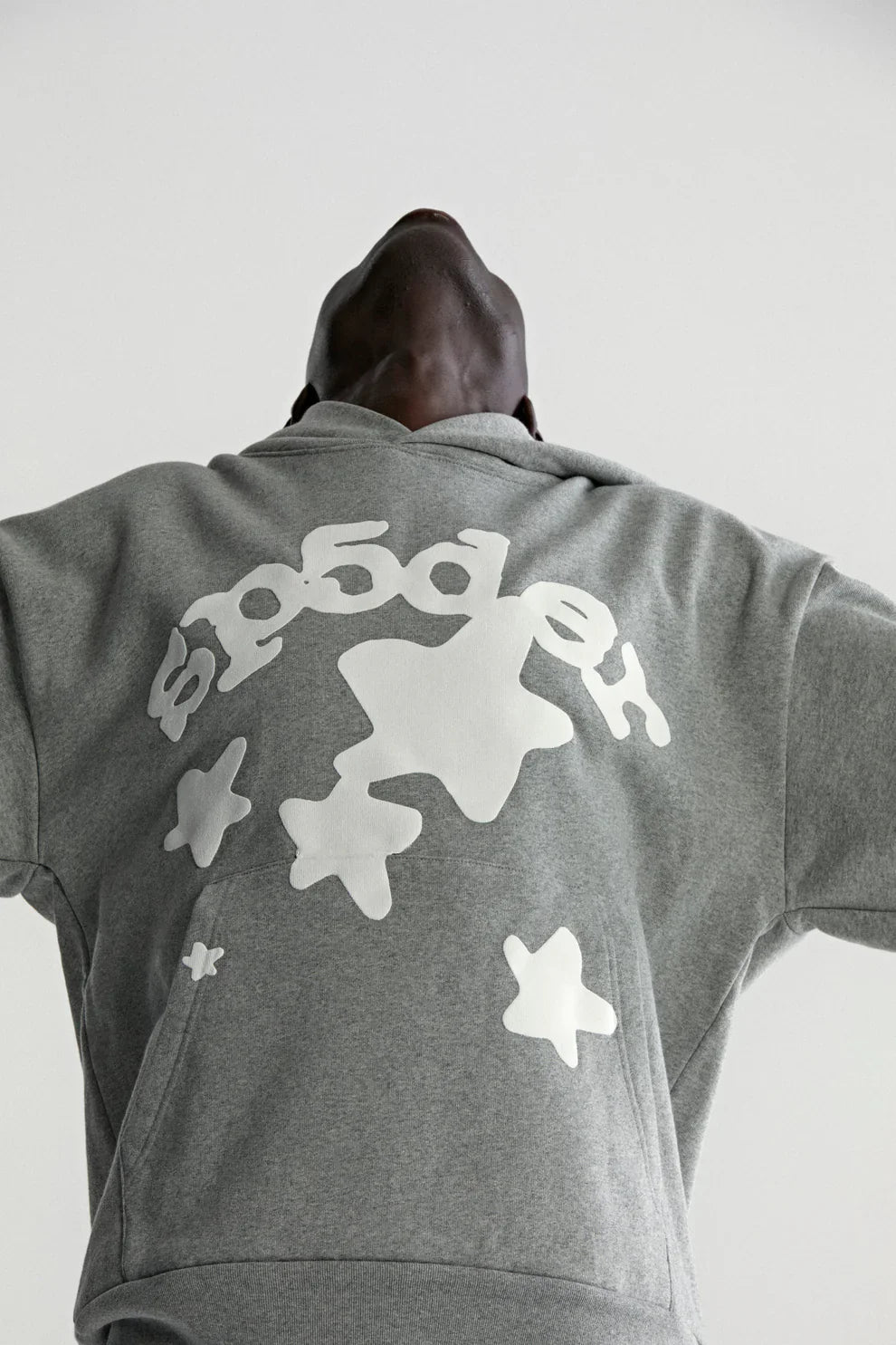 A person wears the Sp5der Stars Hoodie – Grey 440GSM Heavyweight Streetwear Pullover, head tilted back and arms outstretched, viewed from a low angle against a light background.