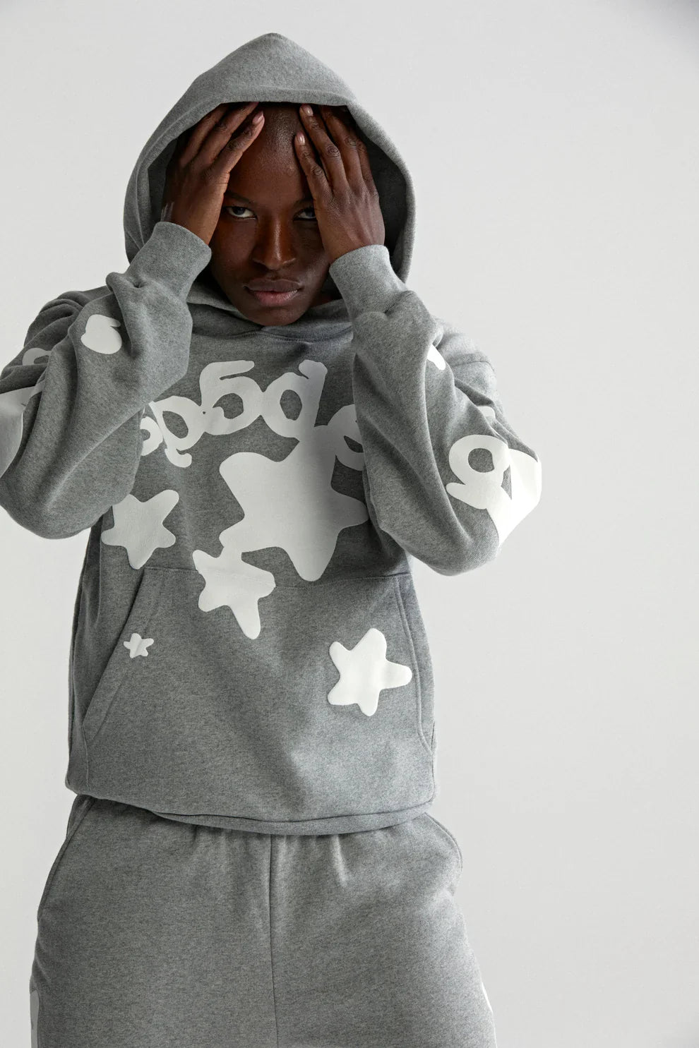 Wearing the Sp5der Stars Hoodie – Grey 440GSM Heavyweight Streetwear Pullover and matching sweatpants with white abstract stars and numbers, a person stands with hands on their head, looking directly at the camera.