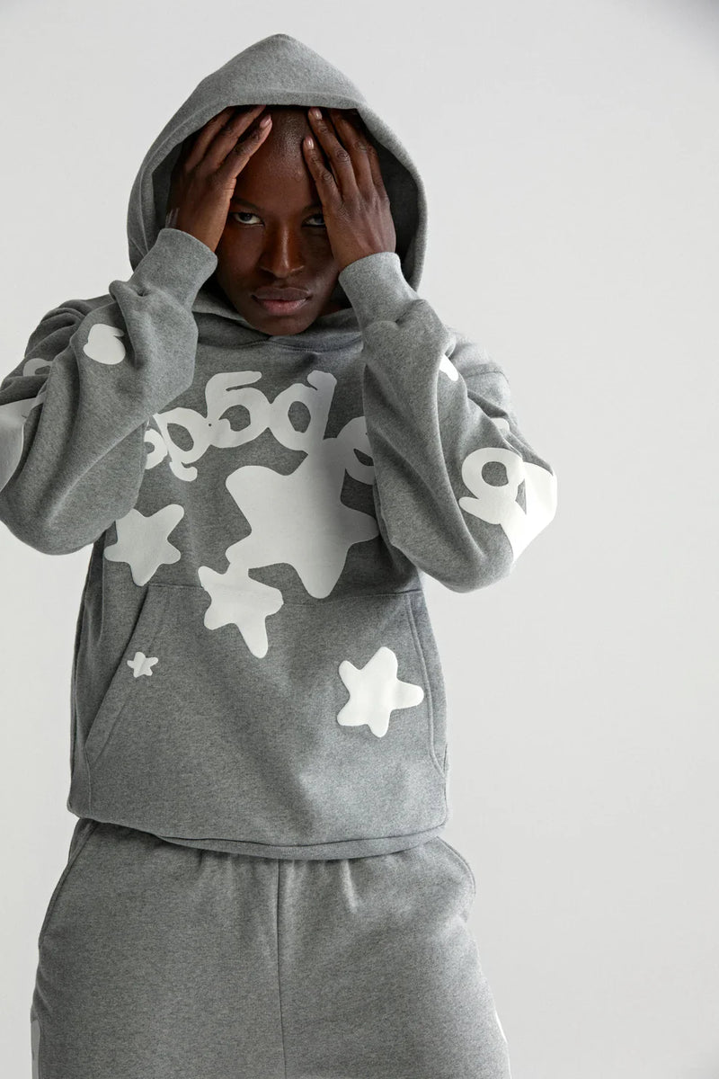 Wearing the Sp5der Stars Hoodie – Grey 440GSM Heavyweight Streetwear Pullover and matching sweatpants with white abstract stars and numbers, a person stands with hands on their head, looking directly at the camera.