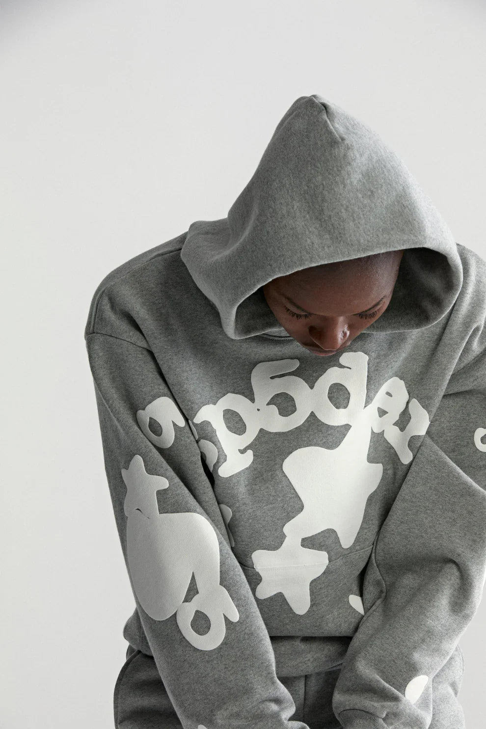 A person in the Sp5der Stars Hoodie – Grey 440GSM Heavyweight Streetwear Pullover, featuring bold white graphics and text, looks downward with the hood up against a plain light background, highlighting its comfort and style.