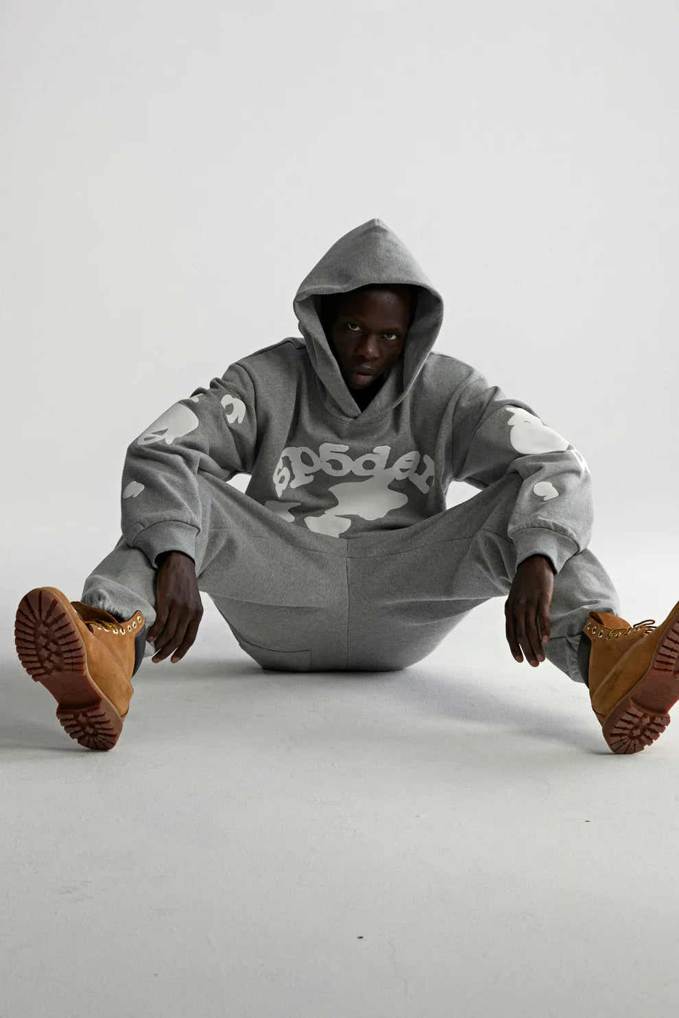 A person in a Sp5der Stars Hoodie – Grey 440GSM Heavyweight Streetwear Pullover with white designs sits on the floor, legs spread and arms on knees, wearing tan boots, looking directly at the camera against a plain white background.