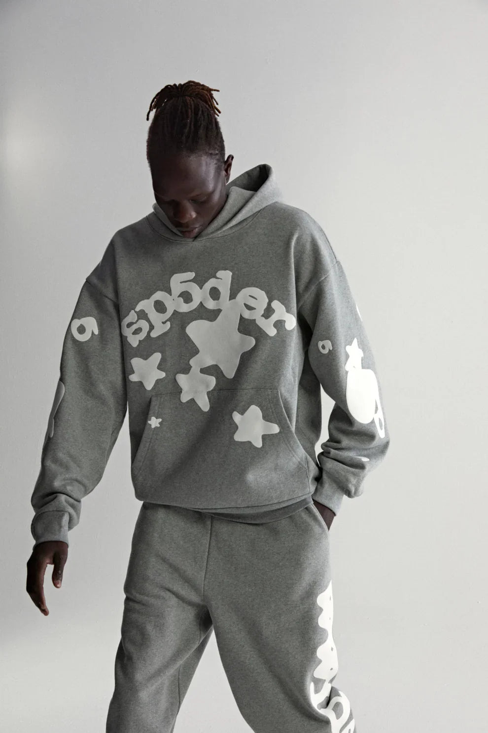 Wearing a Sp5der Stars Hoodie – Grey 440GSM Heavyweight Streetwear Pullover with bold white graphics, a person stands against a plain light gray background, looking down with hands in their pockets.