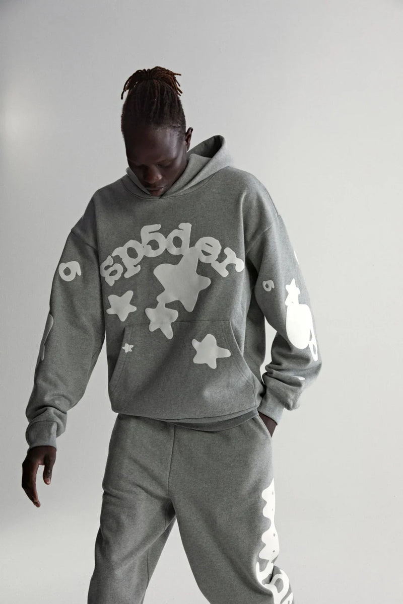 Wearing a Sp5der Stars Hoodie – Grey 440GSM Heavyweight Streetwear Pullover with bold white graphics, a person stands against a plain light gray background, looking down with hands in their pockets.