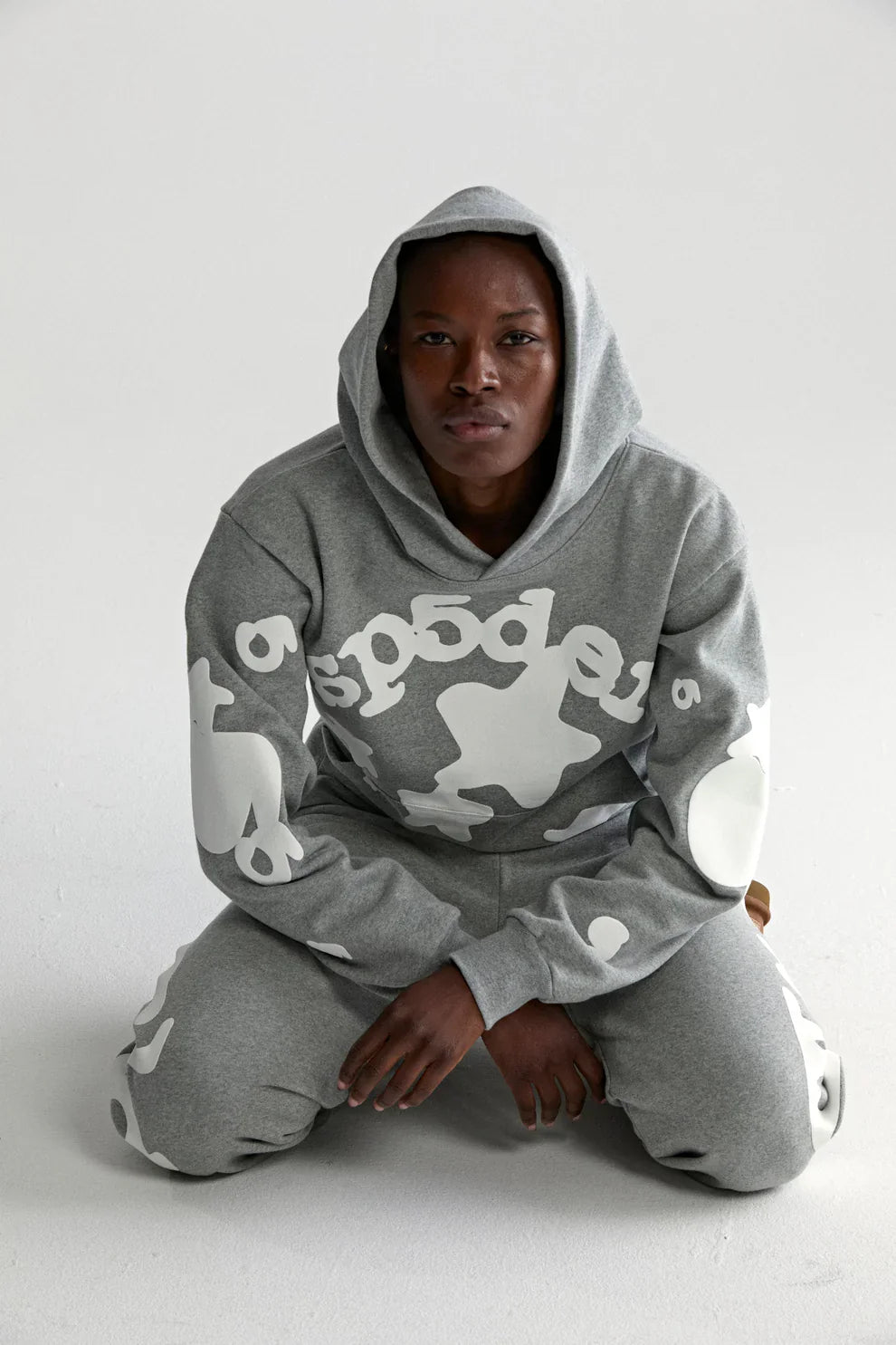 Wearing the Sp5der Stars Hoodie – Grey 440GSM Heavyweight Streetwear Pullover, a person kneels on the floor and looks at the camera; bold white abstract patterns accent the gray heavyweight cotton fleece against a plain background.
