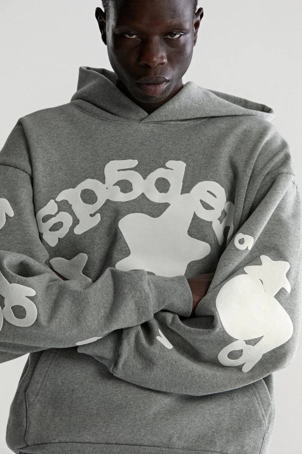 A person wearing the Sp5der Stars Hoodie – Grey 440GSM Heavyweight Streetwear Pullover with bold white graphics stands arms crossed, facing the camera against a plain background. The cotton fleece offers comfort and style.