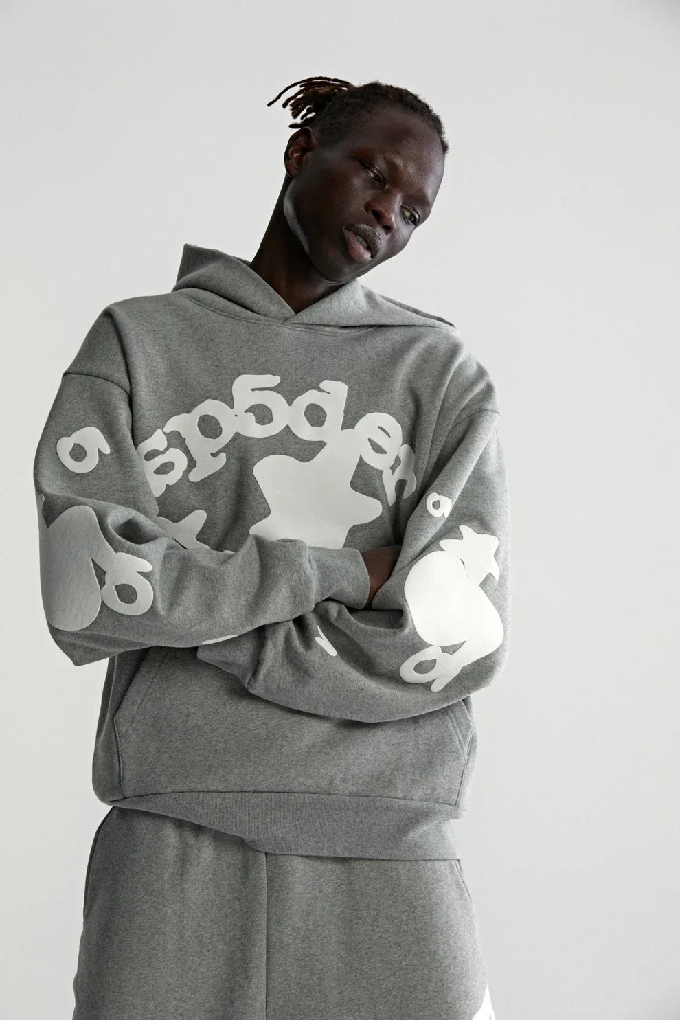 A person with dark skin and short, twisted hair wears the Sp5der Stars Hoodie – Grey 440GSM Heavyweight Streetwear Pullover and matching pants, both showcasing bold white abstract designs, against a plain light backdrop.