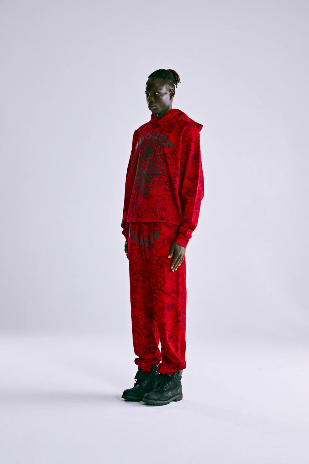 A person stands against a plain background wearing the Red Sp5der Web Hoodie 440GSM with black web graphics, paired with matching red sweatpants and black sneakers.