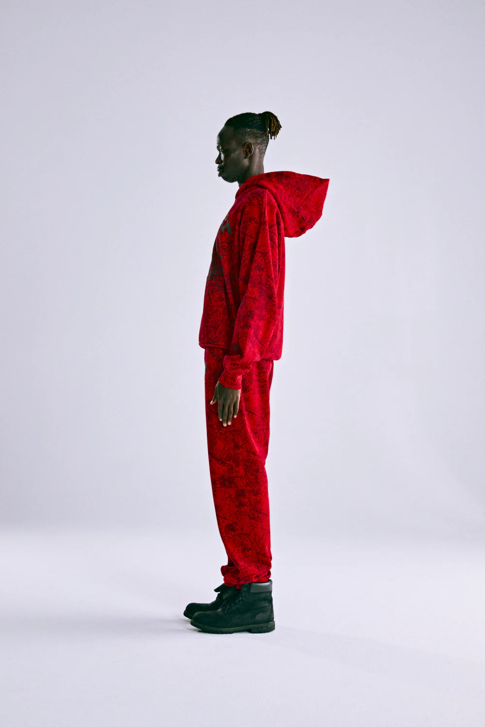 A person stands in profile against a plain background, wearing the Red Sp5der Web Hoodie 440GSM – Red Spider Web Graphic Hoodie with matching sweatpants and black boots. Their hood is down and short twists complete their laid-back streetwear look.