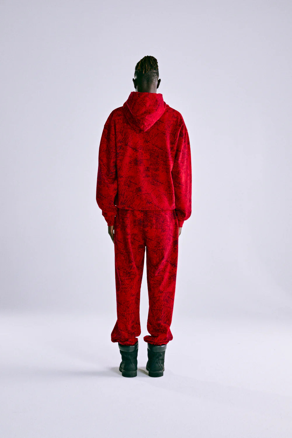 A person stands with their back to the camera, wearing the Red Sp5der Web Hoodie 440GSM – Red Spider Web Graphic Hoodie, paired with matching pants and black boots, against a plain light background.
