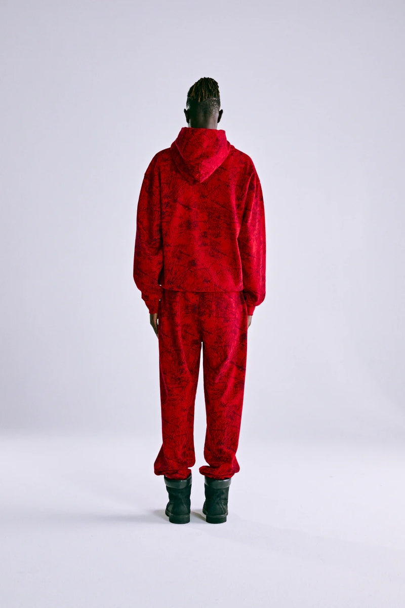 A person stands with their back to the camera, wearing the Red Sp5der Web Hoodie 440GSM – Red Spider Web Graphic Hoodie, paired with matching pants and black boots, against a plain light background.