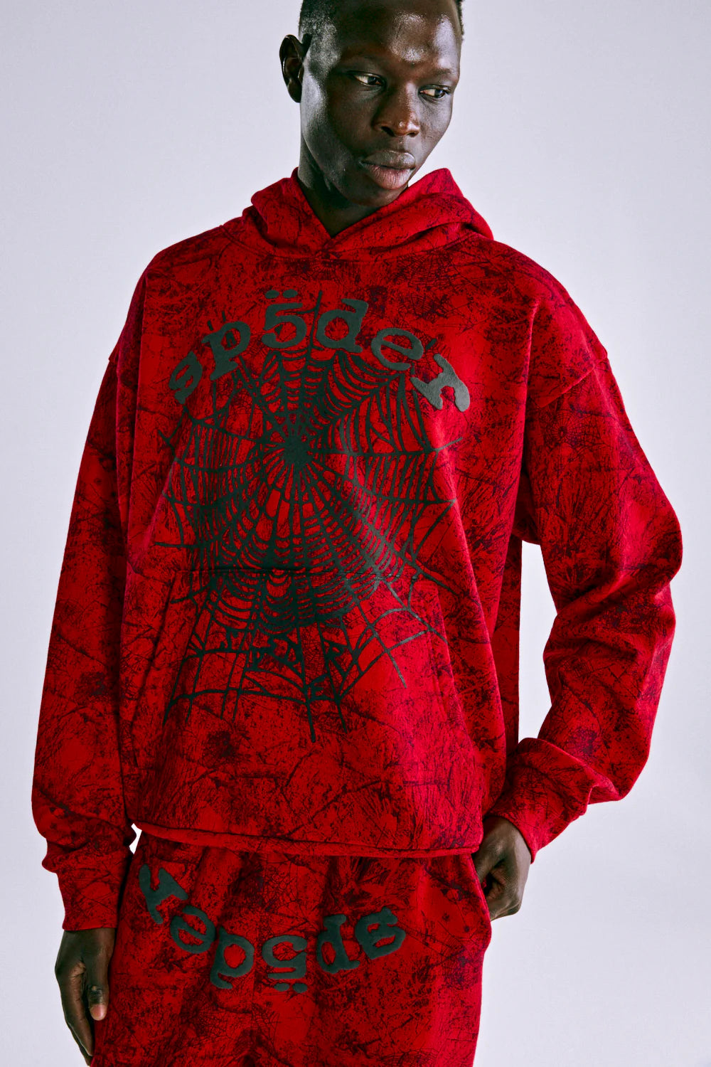 A person wears the Red Sp5der Web Hoodie 440GSM, featuring a bold black spider web and "sp5der" lettering, paired with matching pants, standing against a plain light background.