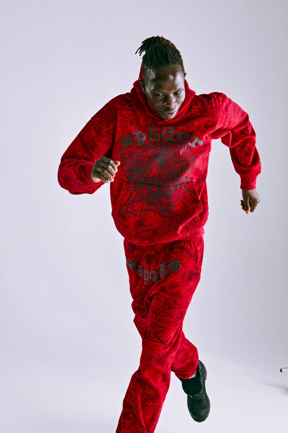 Wearing the Red Sp5der Web Hoodie 440GSM – Red Spider Web Graphic Hoodie and matching sweatpants with bold black accents, a person with dark skin and short twists runs against a plain white background.