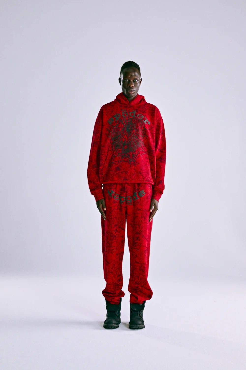 A person faces forward wearing the Red Sp5der Web Hoodie 440GSM – Red Spider Web Graphic Hoodie with matching sweatpants featuring black abstract patterns and black boots, set against a plain light gray background.