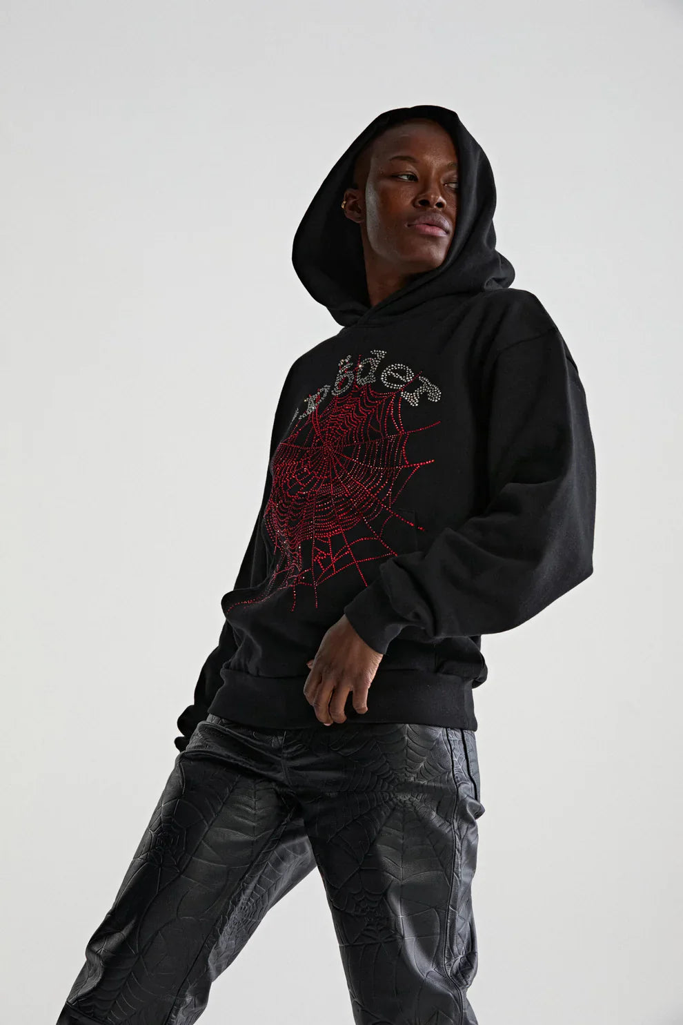 A person wearing the Sp5der Rhinestone Web Hoodie – Black 440GSM Heavyweight Oversized Streetwear Pullover with black textured pants stands against a plain light background, exuding luxury street style while gazing to the side with a hand in their pocket.
