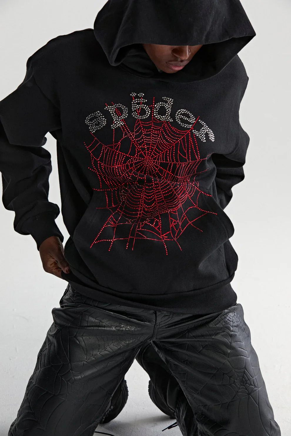 Wearing the Sp5der Rhinestone Web Hoodie – Black 440GSM Heavyweight Oversized Streetwear Pullover, featuring a red rhinestone web and silver “spider,” with textured black pants, kneeling on one knee for luxury streetwear vibes.