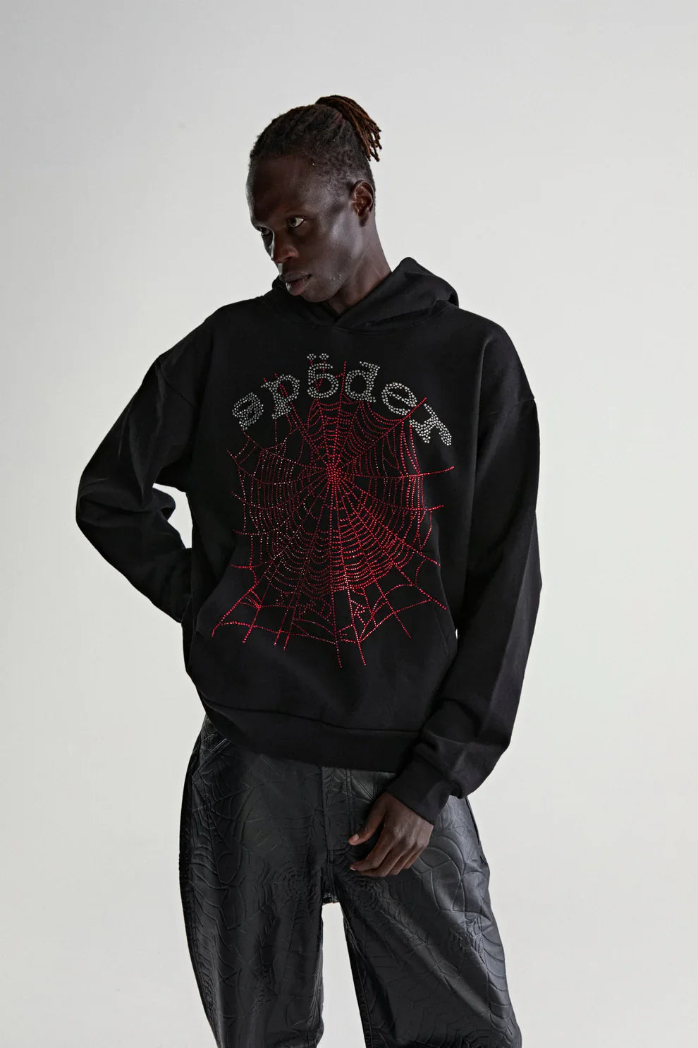 A person in a Sp5der Rhinestone Web Hoodie – Black 440GSM Heavyweight Oversized Streetwear Pullover, with a red spiderweb graphic and stylized "spider" text, stands with one hand on their hip against a light background.