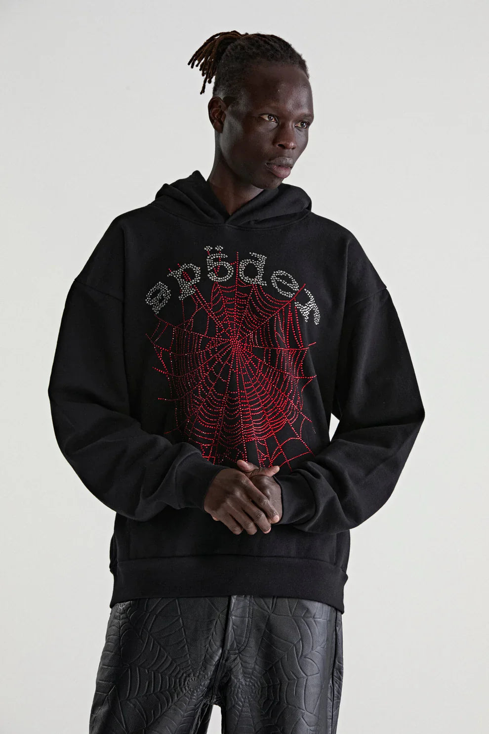 Wearing a Sp5der Rhinestone Web Hoodie – Black 440GSM Heavyweight Oversized Streetwear Pullover with black spiderweb-patterned pants, a person stands before a plain light backdrop, embodying luxury streetwear style.