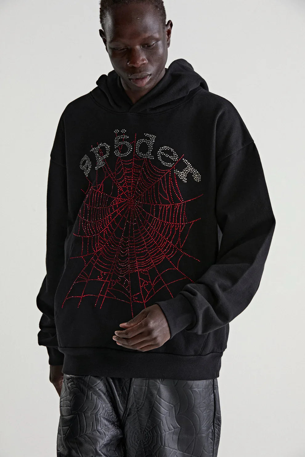 Someone wearing the Sp5der Rhinestone Web Hoodie – Black 440GSM Heavyweight Oversized Streetwear Pullover with red web and stylized "spider" lettering pairs it with dark textured pants, posing against a plain light background for a luxe streetwear look.
