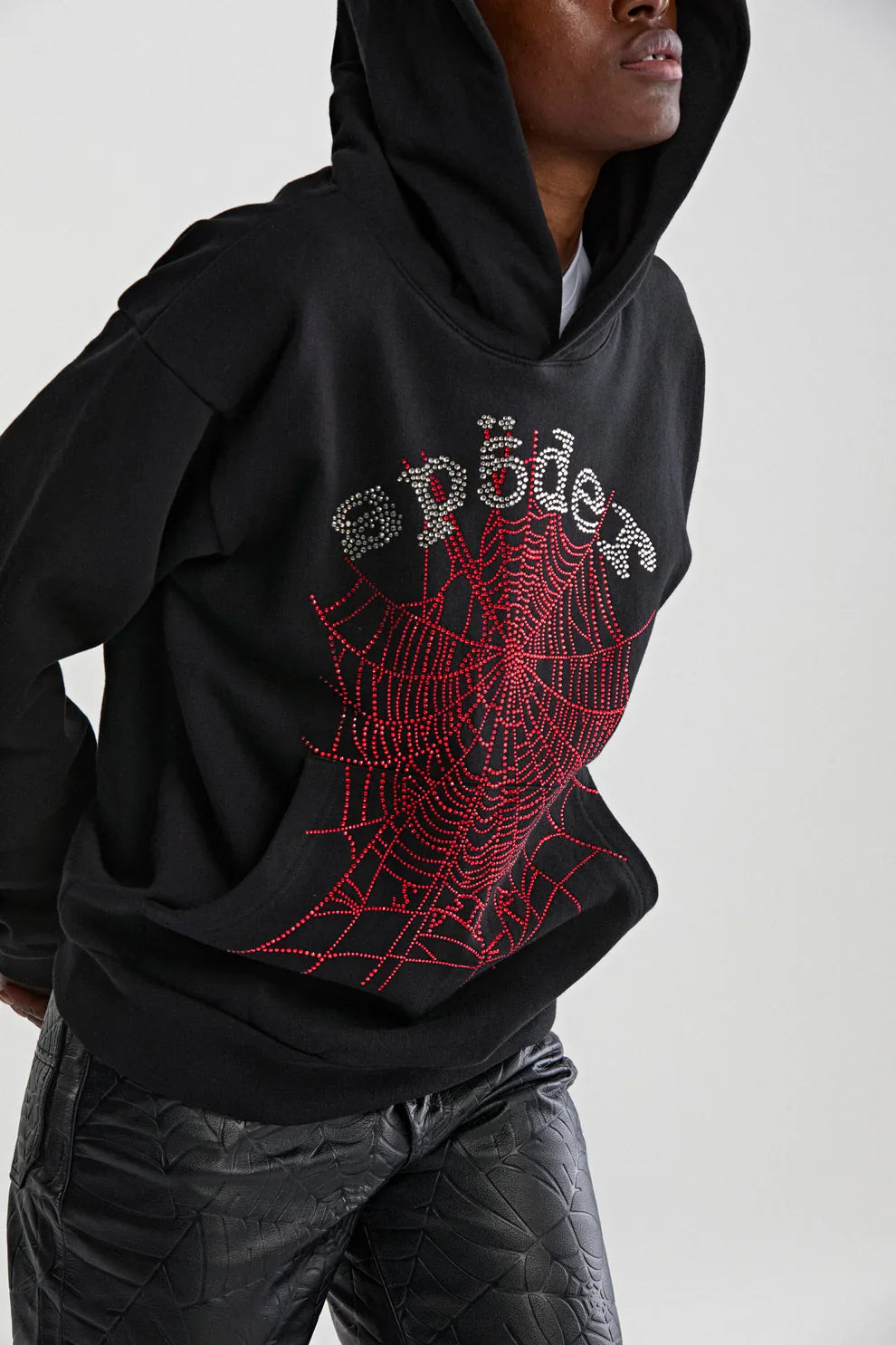 Someone wearing the Sp5der Rhinestone Web Hoodie – Black 440GSM Heavyweight Oversized Streetwear Pullover, featuring a sparkling rhinestone web design, paired with textured black pants for a bold luxury streetwear vibe.