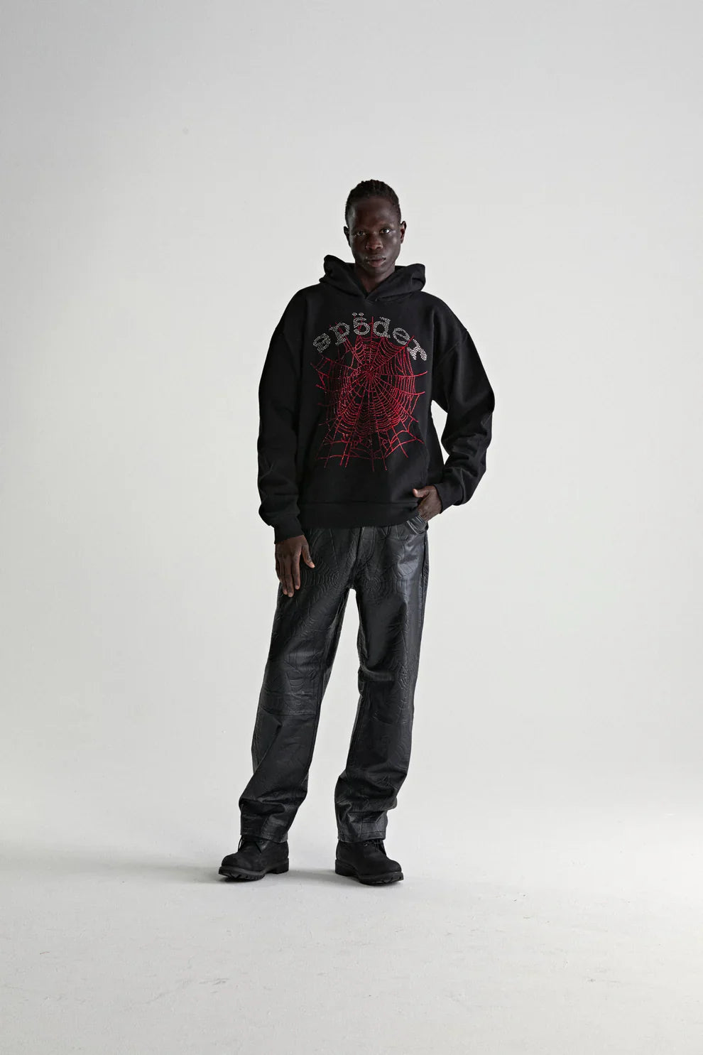 A person stands against a plain background wearing a Sp5der Rhinestone Web Hoodie – Black 440GSM Heavyweight Oversized Streetwear Pullover, paired with black textured pants and shoes for a bold luxury streetwear look.