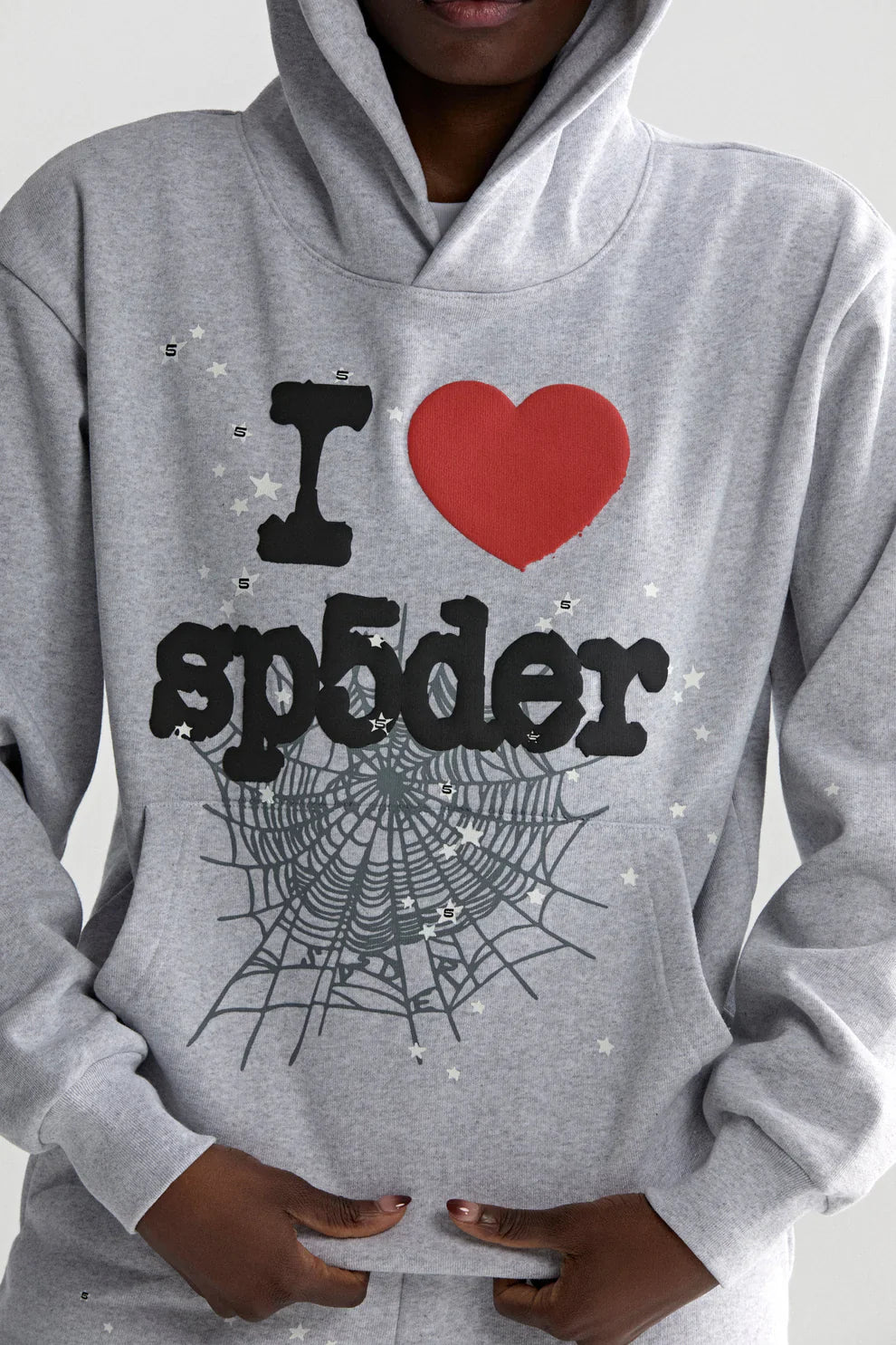 A person sports the Sp5der “I Love Sp5der” Hoodie in gray, a 440GSM heavyweight oversized streetwear pullover with "I ♥ sp5der," a bold red heart, spider web graphics, white stars, and hands tucked in the front pocket.