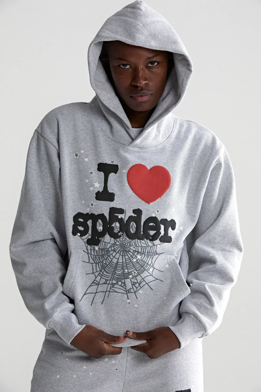 A person in a Sp5der “I Love Sp5der” Hoodie – Grey 440GSM Heavyweight Oversized Streetwear Pullover with “I ♥ sp5der” text and spiderweb graphic stands against a plain light background, facing the camera with hands by the pocket.