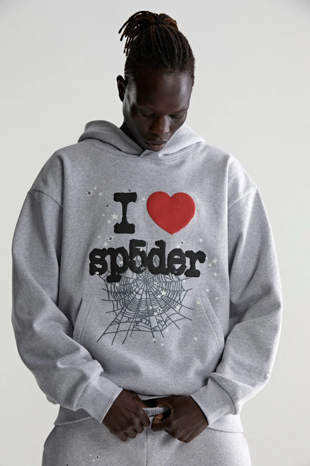A person in a Sp5der “I Love Sp5der” Hoodie – Grey 440GSM Heavyweight Oversized Streetwear Pullover, featuring I ♥ sp5der text and a spider web graphic, channels Y2K style while looking down with hands near their waist.