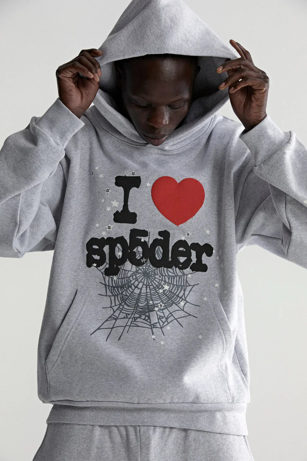 A person wears the Sp5der “I Love Sp5der” Hoodie in grey, a 440GSM heavyweight oversized streetwear pullover featuring “I ♥ sp5der” with a large red heart and spiderweb graphic for a bold Y2K vibe.