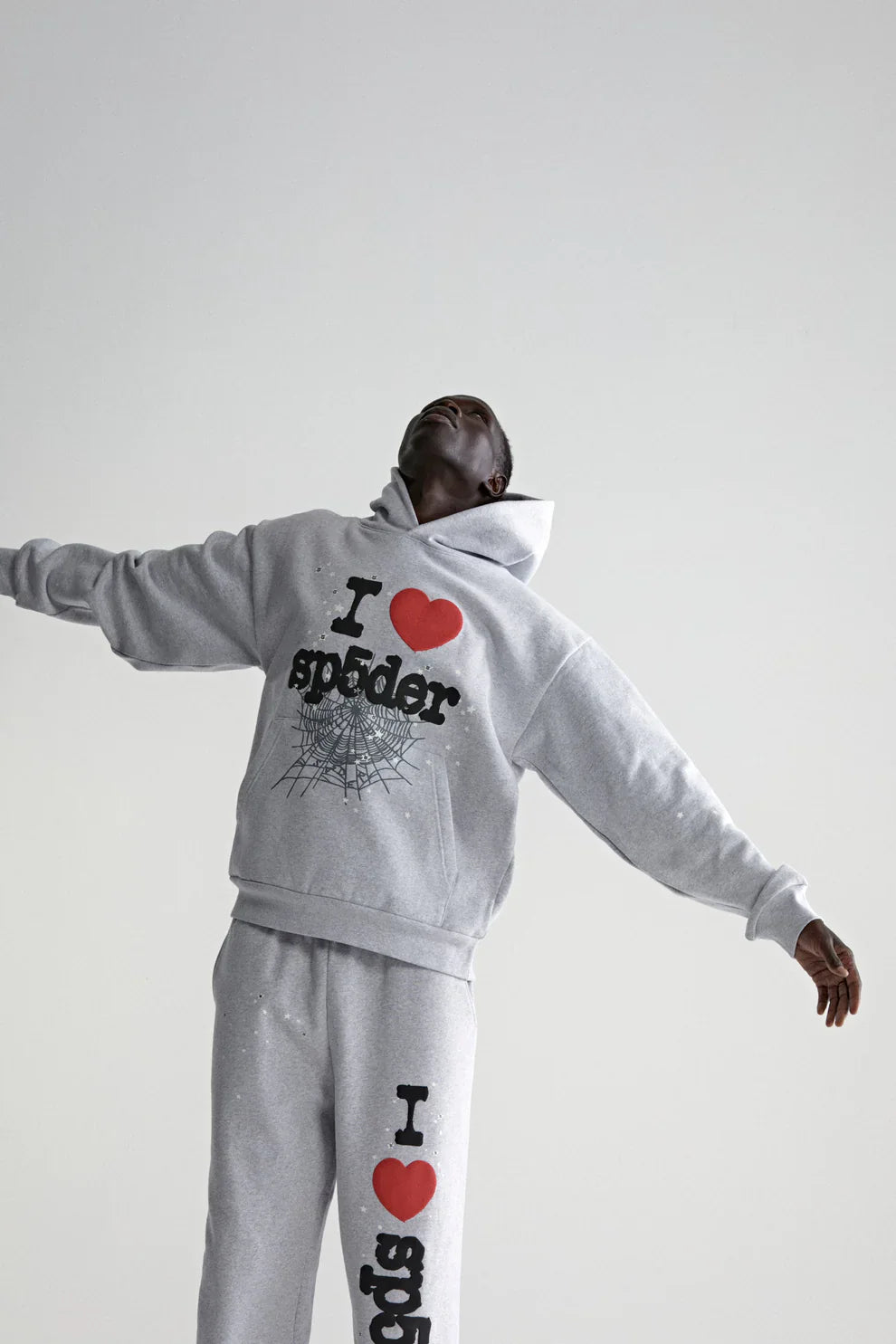 A person stretches their arms wide in a light gray Sp5der “I Love Sp5der” Hoodie—an oversized 440GSM heavyweight cotton fleece pullover with heart and web graphics—showcasing a Y2K streetwear vibe against a plain background.
