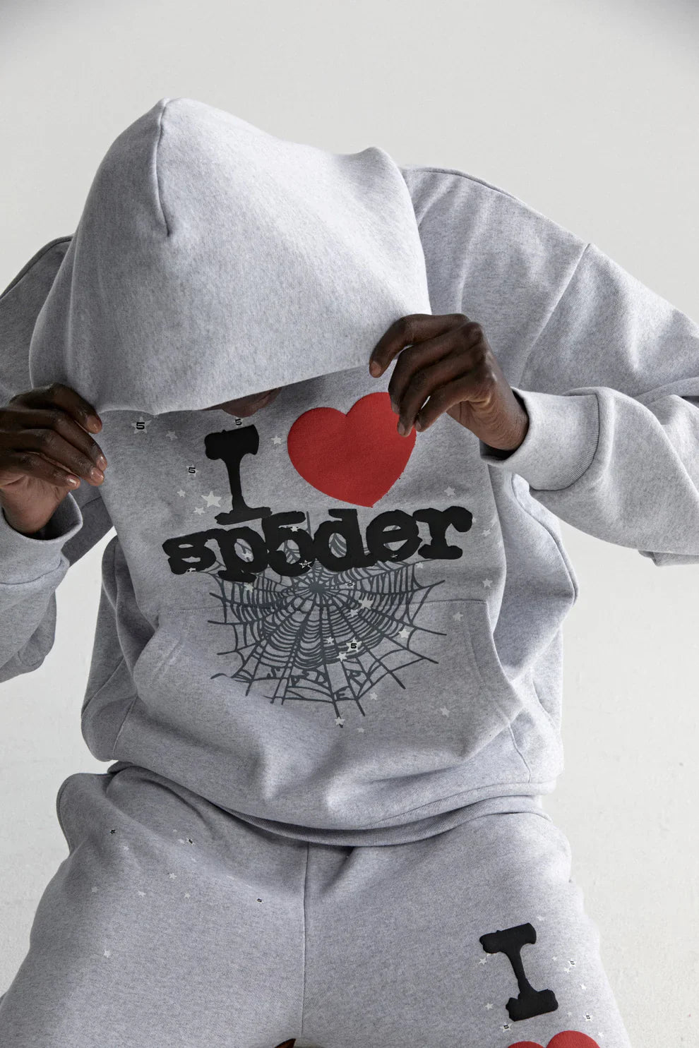 A person wears a grey Sp5der “I Love Sp5der” Hoodie—an oversized 440GSM heavyweight streetwear pullover with I ♥ Sp5der and spiderweb graphics—sitting and pulling the hood over their face against a plain light background.
