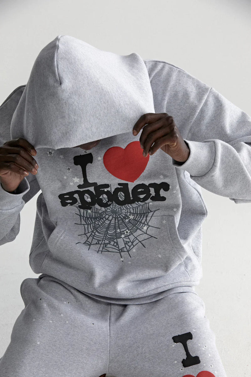 A person wears a grey Sp5der “I Love Sp5der” Hoodie—an oversized 440GSM heavyweight streetwear pullover with I ♥ Sp5der and spiderweb graphics—sitting and pulling the hood over their face against a plain light background.