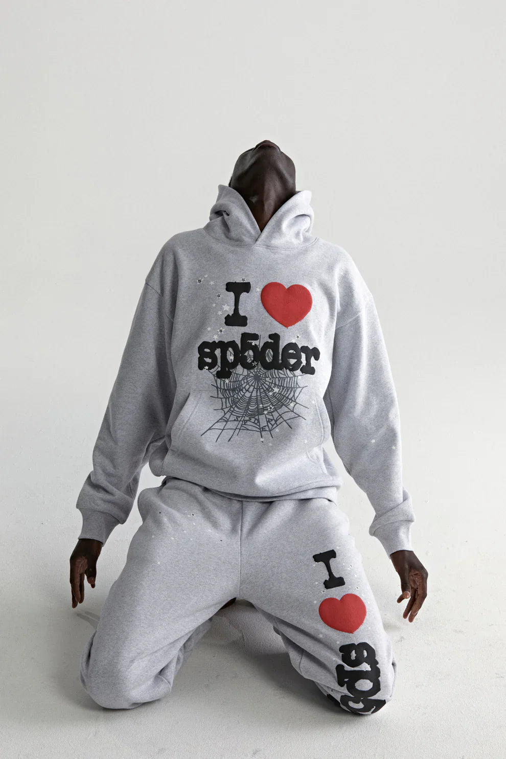A person in a grey Sp5der “I Love Sp5der” Hoodie—440GSM heavyweight, oversized streetwear pullover—kneels and leans back against a white background, evoking Y2K style.