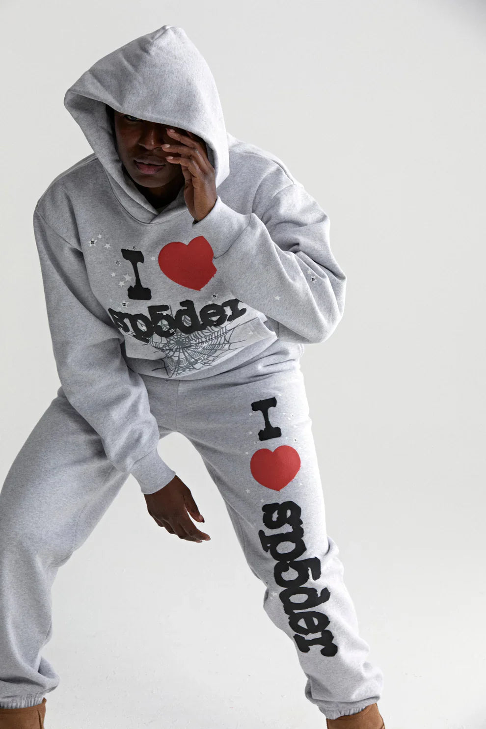 A person in a Sp5der “I Love Sp5der” Grey 440GSM Heavyweight Oversized Streetwear Pullover poses with one hand partly covering their face, showing off the web graphic and I ♥ spider text for a Y2K streetwear vibe.