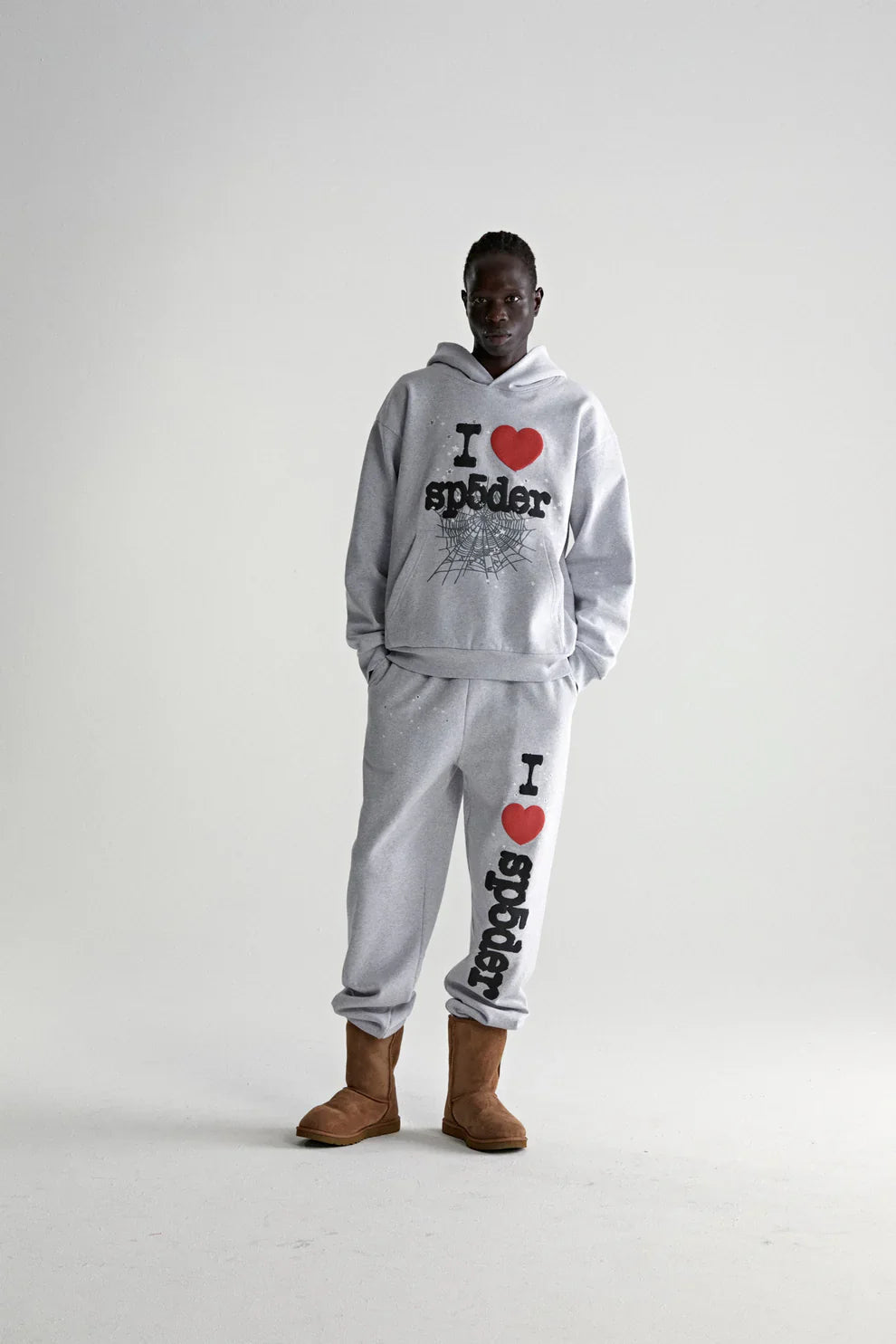 A person stands against a plain background wearing a Sp5der “I Love Sp5der” Hoodie – Grey 440GSM Heavyweight Oversized Streetwear Pullover with matching sweatpants, displaying bold I ❤️ SP5DER text, and paired with brown fuzzy boots for a cozy Y2K look.