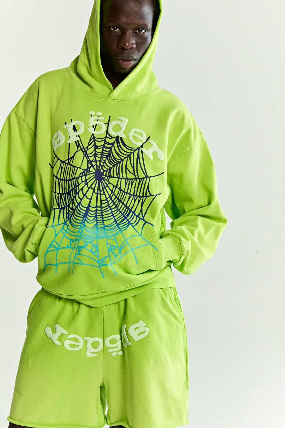 Wearing the Sp5der Web Graphic Hoodie – Neon Green 440GSM Heavyweight Streetwear and matching shorts, a person stands against a plain light background. The hoodie features a bold black spider web graphic and "spider" in white.