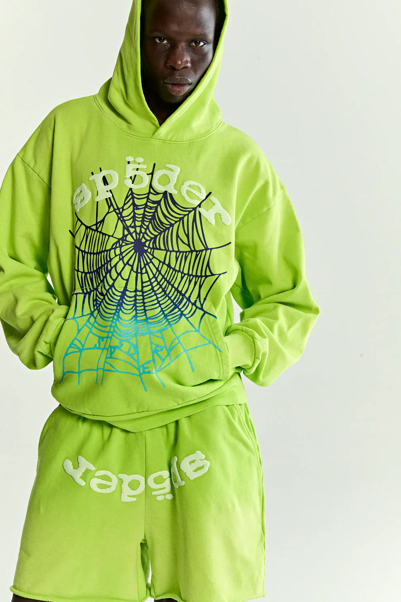 Wearing the Sp5der Web Graphic Hoodie – Neon Green 440GSM Heavyweight Streetwear and matching shorts, a person stands against a plain light background. The hoodie features a bold black spider web graphic and "spider" in white.