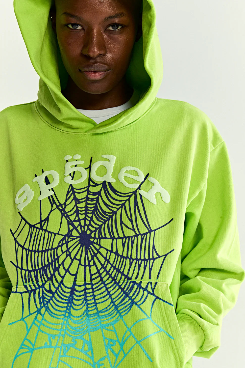 A person wears the Sp5der Web Graphic Hoodie – Neon Green 440GSM Heavyweight Streetwear, featuring a bold blue and black spiderweb and white "sp5der" text on the chest, with the hood up against a plain white background.